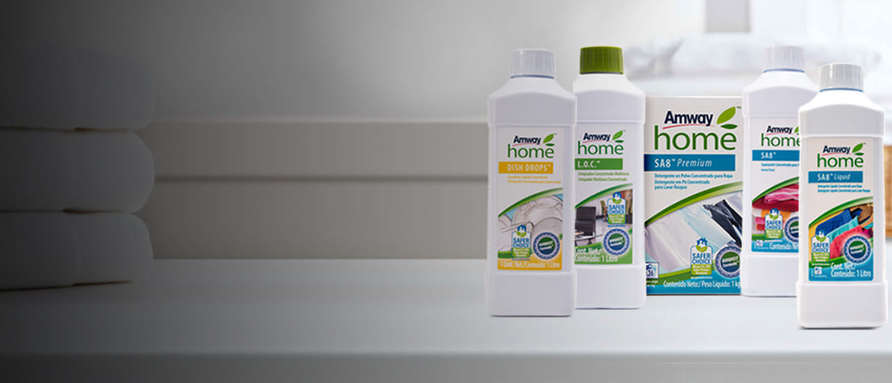 Imagen con varios productos de limpieza de Amway HomeT. Detrás de ellos hay dos pilas de tres toallas plegadas. Productos de Limpieza Amway Home, seguros y efectivos. Conoce los ingredientes de estos productos derivados de la naturaleza.