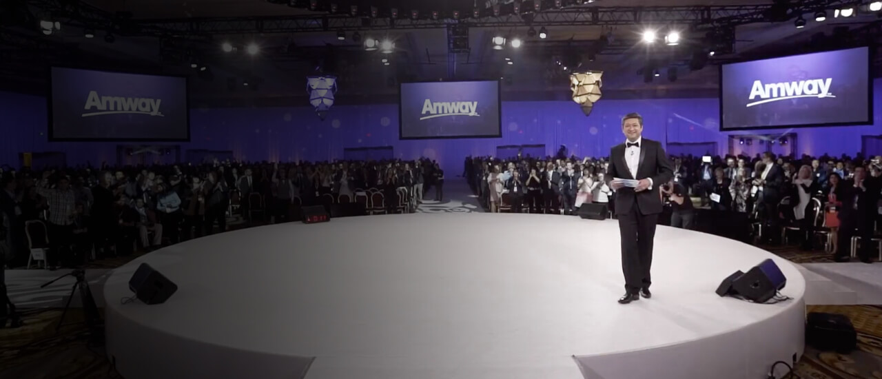 Imagen de un escenario en un evento donde se otorgan reconocimientos, en él, sobre una plataforma blanca, hay un hombre vestido con smoking. Hay público en el fondo. En Amway reconocemos tus logros.